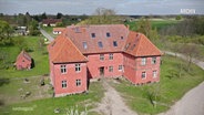 Ein markantes, rotes Herrenhaus auf einer Wiese. © Screenshot 