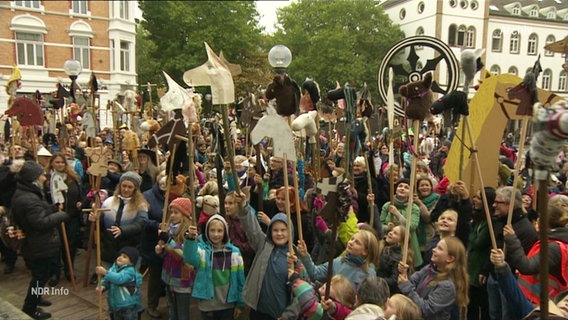 Eine Menge Kinder steht auf einem Marktplatz und streckt Steckenpferde in die Höhe. © Screenshot 