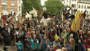 Eine Menge Kinder steht auf einem Marktplatz und streckt Steckenpferde in die Höhe. © Screenshot 