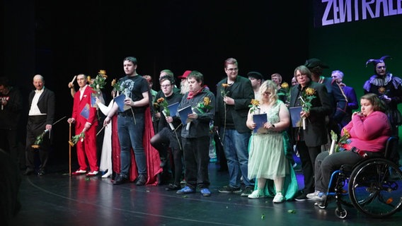 Das Esemble der Theatergruppe "Die Eckigen" steht mit Blumen in den Händen auf der Bühne. © Screenshot 