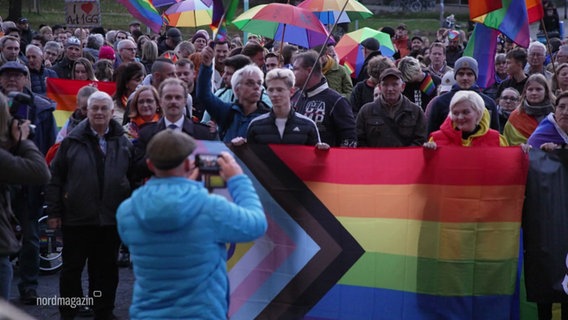 Menschen stehen mit bunten Schirmen und Regenbogenflaggen vor dem Neubrandenburger Rathaus. © Screenshot 