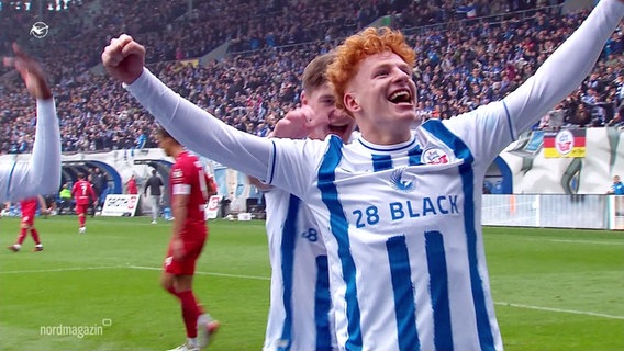 Hansa-Rostock-Spieler Tim Krohn bei einem Torjubel. Er trägt ein weißes Trikot mit blauen Streifen, hat rote zersauste Haare und reißt die Arme zum Jubeln in die Luft. © Screenshot 