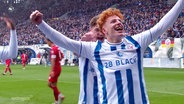 Hansa-Rostock-Spieler Tim Krohn bei einem Torjubel. Er trägt ein weißes Trikot mit blauen Streifen, hat rote zersauste Haare und reißt die Arme zum Jubeln in die Luft. © Screenshot 