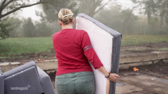 Die Kamera folgt einer Frau, die ein Teil eines Sofas trägt und es offenbar zu weiteren Teilen auf einem Recyclinghof legen möchte. © Screenshot 