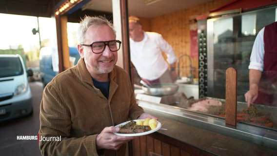 Kochbuch-Autor Stefan Paul grinsend mit einem Teller Grünkohl in der Hand vor einem Streetfood-Stand © Screenshot 