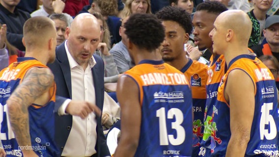 Ein Basketball-Coach bespricht sich mit seinen Spielern bei einem Spiel. © Screenshot 