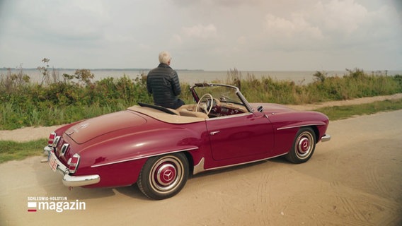 Ein Mann lehnt an einem roten Oldtimer und blickt aufs Meer. © Screenshot 