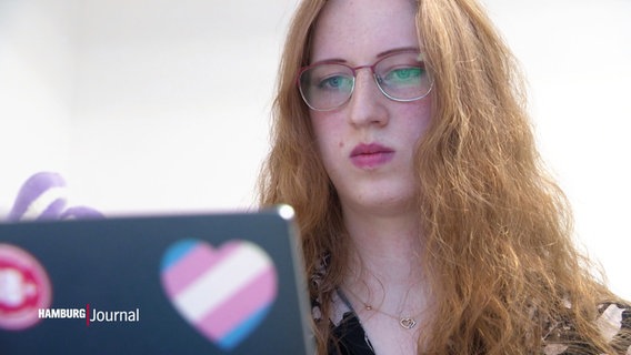 Eine Frau sitzt konzentriert vor einem Laptop auf dessen Rückseite eine Herz-Sticker in den Symbol-Farben der Transgender-Community klebt. © Screenshot 