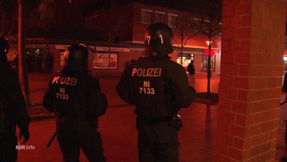 Zwei Einsatzkräfte der Polizei beobachten in Schutzausrüstung jugendliche Randalierende auf einem Platz bei Nacht. © Screenshot 