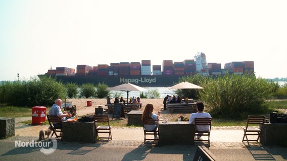 Vor einem Strandcafé an der Elbe fährt ein riesiges Container-Schiff vorbei. © Screenshot 