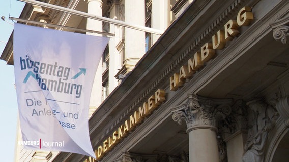 Blick auf die Fassade der Handelskammer in Hamburg. Über Säulen formen goldene Buchstaben die Worte "Handelskammer Hamburg". Davor ist eine Flagge befestigt, die den Börsentag bewirbt. © Screenshot 