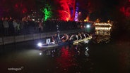 Ein Team startet mit seinem Boot beim Lichterpaddeln auf dem Neubrandenburger Oberbach, im Hintergrund bunt angestrahlte Bäume. © Screenshot 