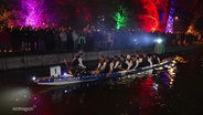 Ein Drachenboot-Team macht sich unter den Augen vieler Schaulustiger zum Start bereit. Boot und Ufer sind bunt beleuchtet. © Screenshot 