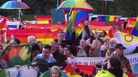 Viele Menschen stehen mit Regenbogenflaggen und Regenbogensymbolen zusammen. © Screenshot 