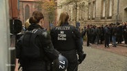 Polizistinnen beobachten eine Gruppe von Menschen vor einem Gebäude. © Screenshot 