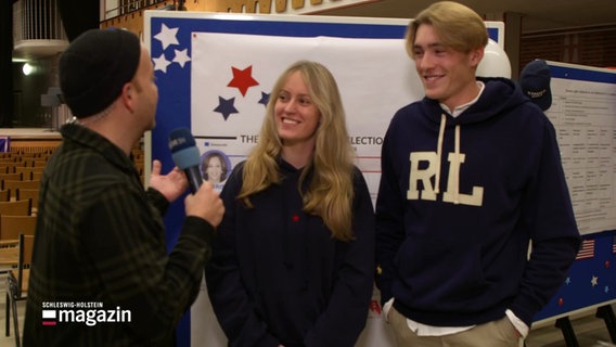 Ein Reporter interviewt zwei Menschen vor einer Tafel mit Informationen zur US-Präsidentschaftswahl. © Screenshot 