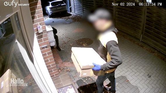 Bild einer Überwachungskamera: Ein als Paketbote verkleideter Mann steht vor einer Wohnungstür, eine weitere, dunkel gekleidete Person versteckt sich neben der Tür. © Screenshot 