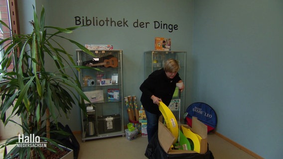 Eine Frau sortiert in einem Raum Gegenstände ein, an der Wand steht "Bibliothek der Dinge". © Screenshot 