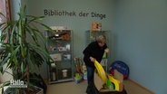 Eine Frau sortiert in einem Raum Gegenstände ein, an der Wand steht "Bibliothek der Dinge". © Screenshot 