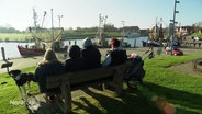 Touristen in Greetsiel sitzen auf einer Bank am Hafen. © Screenshot 