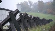 Kreuze liegen in einer Reihe auf einem Acker vor einer Mauer, im Hintergrund ein Grenzturm. © Screenshot 