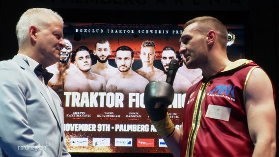 Boxer Denis Radovan mit Mikrofon, er spricht mit einem Mann mit weißem Sakko und Fliege, im Hintergrund ein Plakat der "Traktor Fight Night 1". © Screenshot 