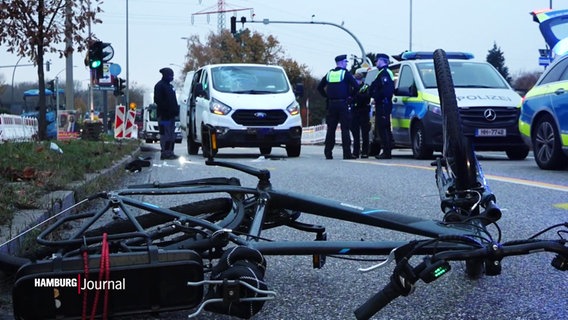 Ein Fahrrad liegt an der Unfallstelle auf der Straße. Im Hintergrund stehen Polizeibeamte und -Fahrzeuge. © Screenshot 