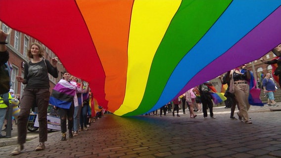 Menschen ziehen mit einer langen Regenbogenfahne durch eine Innenstadt. © Screenshot 