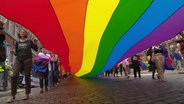 Menschen ziehen mit einer langen Regenbogenfahne durch eine Innenstadt. © Screenshot 