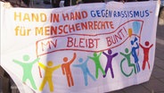 Mit einem bunten Spruchbanner demonstrieren mehrere Personen für Vielfalt, Menschenrechte und gegen Rassismus. © Screenshot 