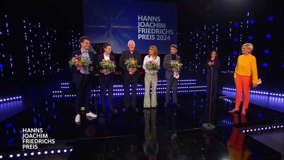 Die Preisträger*innen des Hanns-Joachim-Friedrichs-Preis 2024 (v.l.n.r.): Lutz van der Horst, Fabian Köster, Jan Lorenzen, Eva Schulz und Paul Schwemm. Sie stehen in einer Reihe und halten je einen Blumenstrauß in der Hand. Neben ihnen steht Moderatorin Susanne Stichler. © Screenshot 