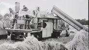 Ein historisches Schwarz-Weiß-Foto zeigt Feldarbeiter auf einer größeren Dieselmaschine für die landwirtschaftliche Arbeit. © Screenshot 