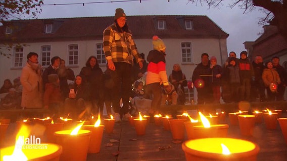 Viele Kerzen in Gefäßen, die vor Wind schützen sollen, stehen auf einem Boden. Im Hintergrund sind Menschen in wintertauglicher Kleidung. © Screenshot 