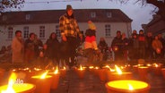 Viele Kerzen in Gefäßen, die vor Wind schützen sollen, stehen auf einem Boden. Im Hintergrund sind Menschen in wintertauglicher Kleidung. © Screenshot 