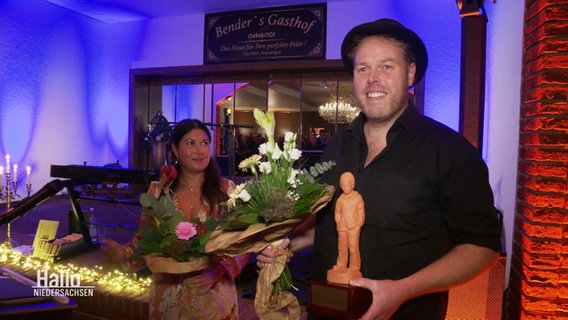 Preisträger Frank Jakobs. Ein hochgewachsener Mann, mit einem hellen Dreitagebart und einem schwarzen Hut auf dem Kopf. Er hält einen Blumenstrauß und den Keerlke-Preis in den den Händen. © Screenshot 