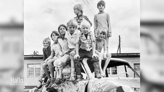 Eine schwarz,weiße Fotografie: Jungen im Grundschulalter und ausgetragenen Klamotten, stehen in der Gruppe auf dem Wrack eines alten Autos. © Screenshot 