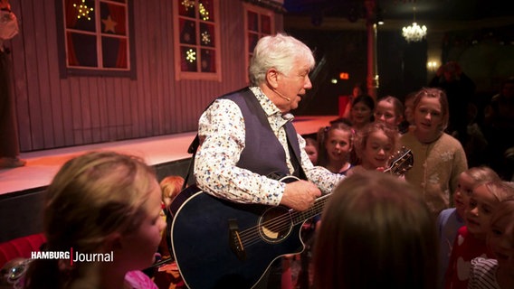 Kinderliedersänger Rolf Zukowski steht mit einer Gitarre, singend und von Kindern umringt im Schmidt's Tivoli © Screenshot 