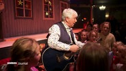 Kinderliedersänger Rolf Zukowski steht mit einer Gitarre, singend und von Kindern umringt im Schmidt's Tivoli © Screenshot 