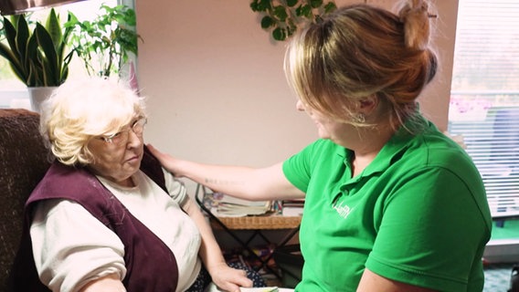 Eine Pflegekraft sitzt bei einer Patientin zuhause und streichelt deren Schulter. © Screenshot 