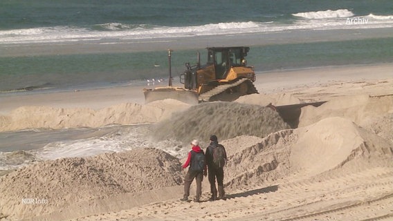 Mit einer sogenannten Raupe wird frischer Sand an einem Strand verteilt. Zwei Passanten sehen dabei zu. © Screenshot 