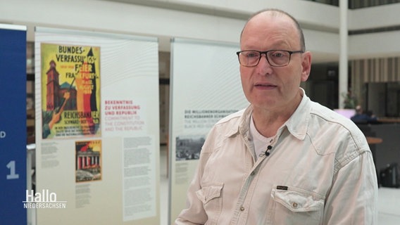 Ralf Hermes, Regionalsprecher Hannover des Reichsbanner Schwarz-Rot-Gold, steht vor den im Landtag ausgestellten Plakaten zum 100-jährigen Jubiläum des Reichsbanners. © Screenshot 