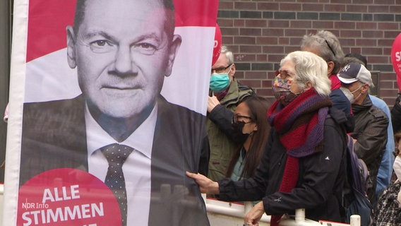 Eine Frau zieht an einem Werbebanner mit dem Konterfei von Olaf Scholz. © Screenshot 