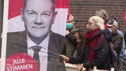 Eine Frau zieht an einem Werbebanner mit dem Konterfei von Olaf Scholz. © Screenshot 