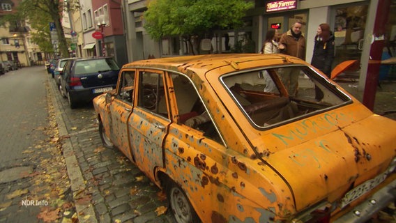 Ein zerschossenes orangenes Auto mit ukrainischem Kennzeichen steht auf einem Parkplatz am Straßenrand. © Screenshot 