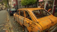 Ein zerschossenes orangenes Auto mit ukrainischem Kennzeichen steht auf einem Parkplatz am Straßenrand. © Screenshot 