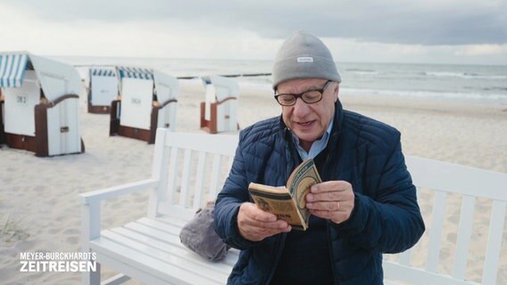 Hubertus Meyer-Burckhardt liest auf einer Bank am Strand aus einem historischen Reiseführer vor. © Screenshot 