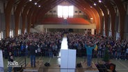 Ein Chor mit 1000 Sänger:innen bei den Proben in einer großen Halle. © Screenshot 