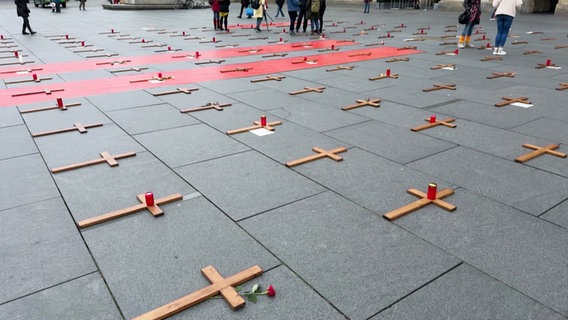 Holzkreuze liegen auf einem öffentlichen Platz, daneben teilweise rote Grabkerzen oder Rosen. © Screenshot 