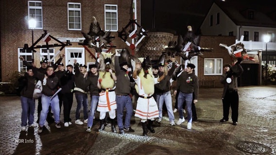 Gruppenbild: Mit Schaffellen und Federn verkleidete, jubelnde Personen, es werden selbst gezimmerte Objekte in die Luft gehalten. © Screenshot 