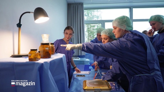 Schülerinnen und Schüler in OP-Kleidung bei einem Workshop in einer Klinik. Eine Person zeigt auf Präparate in braunen Gläsern. © Screenshot 